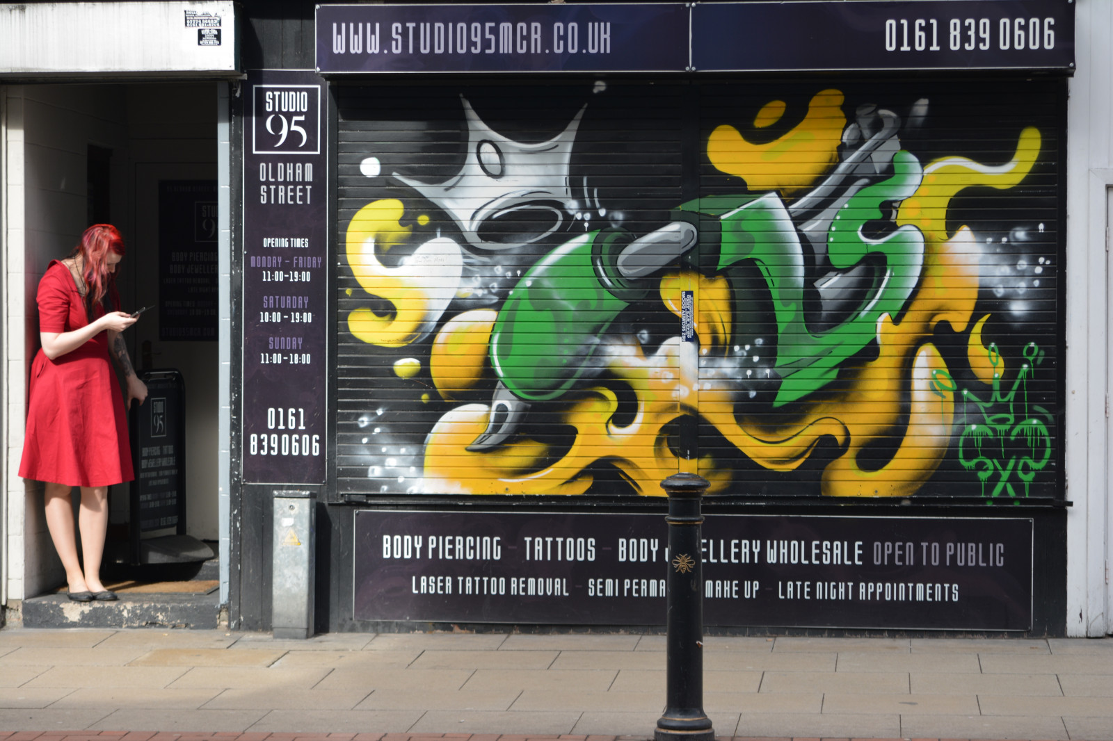 England, Großbritannien, Manchester, Manchesterstreets, Strassenfotografie, Streetscene, Urbanscen, Urbanart, Straßenkunst, Kunstwerk, Kunst, Rollläden, Shuttermural, Wandmalereien, Bürgersteig, Pflaster, draussen, Schaufenster, Geschäft, Tattoopalour, Studio95, Mädchen, weiblich, Frau, Tür, Texting, Poller, Tätowierungen, rotes Kleid, Oldhamstreet, Nördliches Viertel, Rote Haare, Girlinreddress, M4